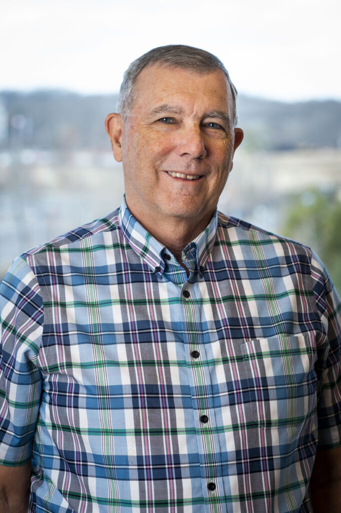 Phil Arkin, wearing a plaid shirt, smiles in front of a blurred landscape