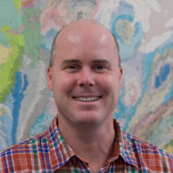 James Farquhar smiles for the camera, wearing a red and blue gingham button-up in front of a map-like abstract background.