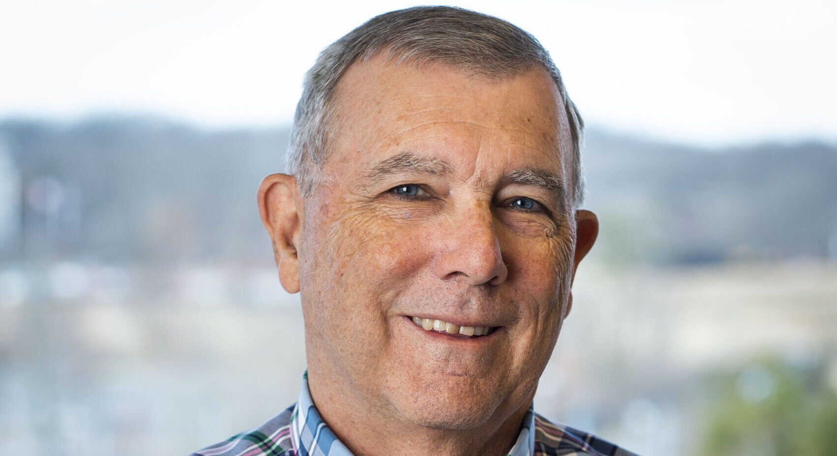 Phil Arkin, wearing a plaid shirt, smiles in front of a blurred landscape