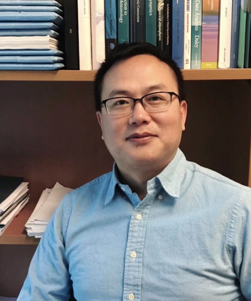 Jifu Yin poses in front of his bookshelf, wearing a blue button-down