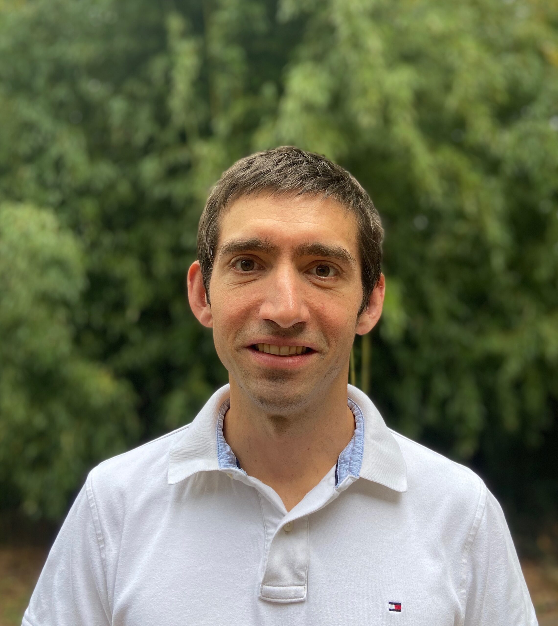 Dr. Muncak smiles in front of a woodsy landscape, wearing a white polo shirt