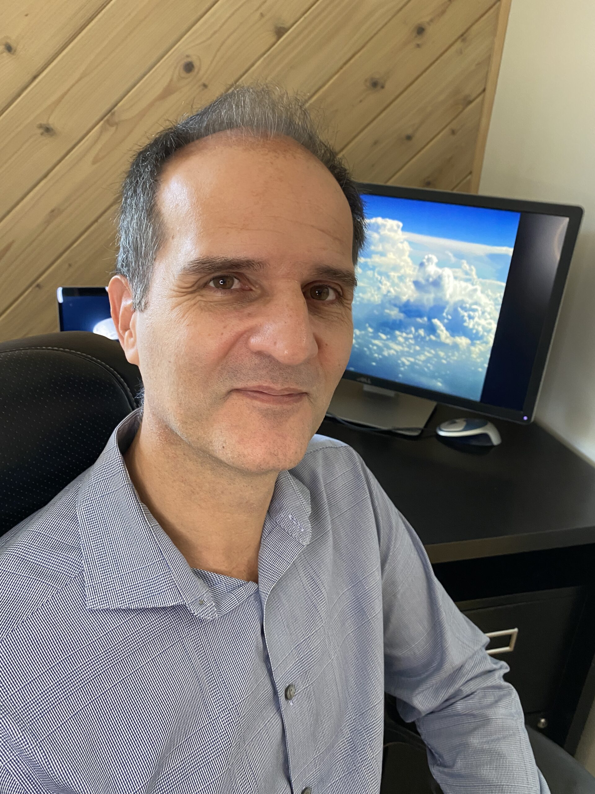 Dr. Kollias sits in his home office, smiling at the camera, and wearing a blue button-down shirt
