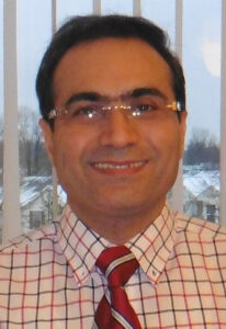 Isaac Moradi, wearing rimless glasses and a red shirt and tie, smiles at the camera
