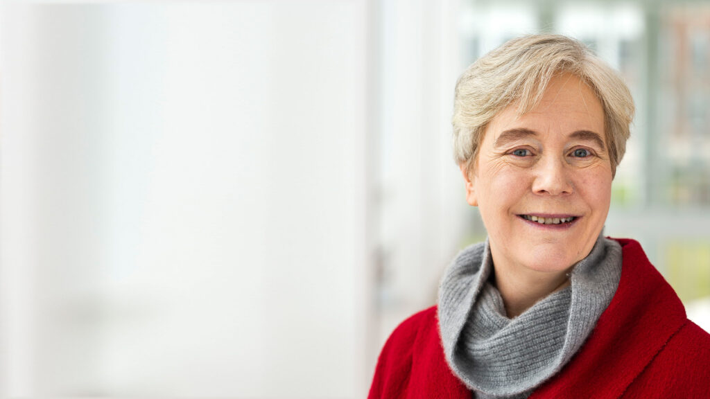 Ellen Williams, wearing a red coat and a grey scarf, smiles for the camera.