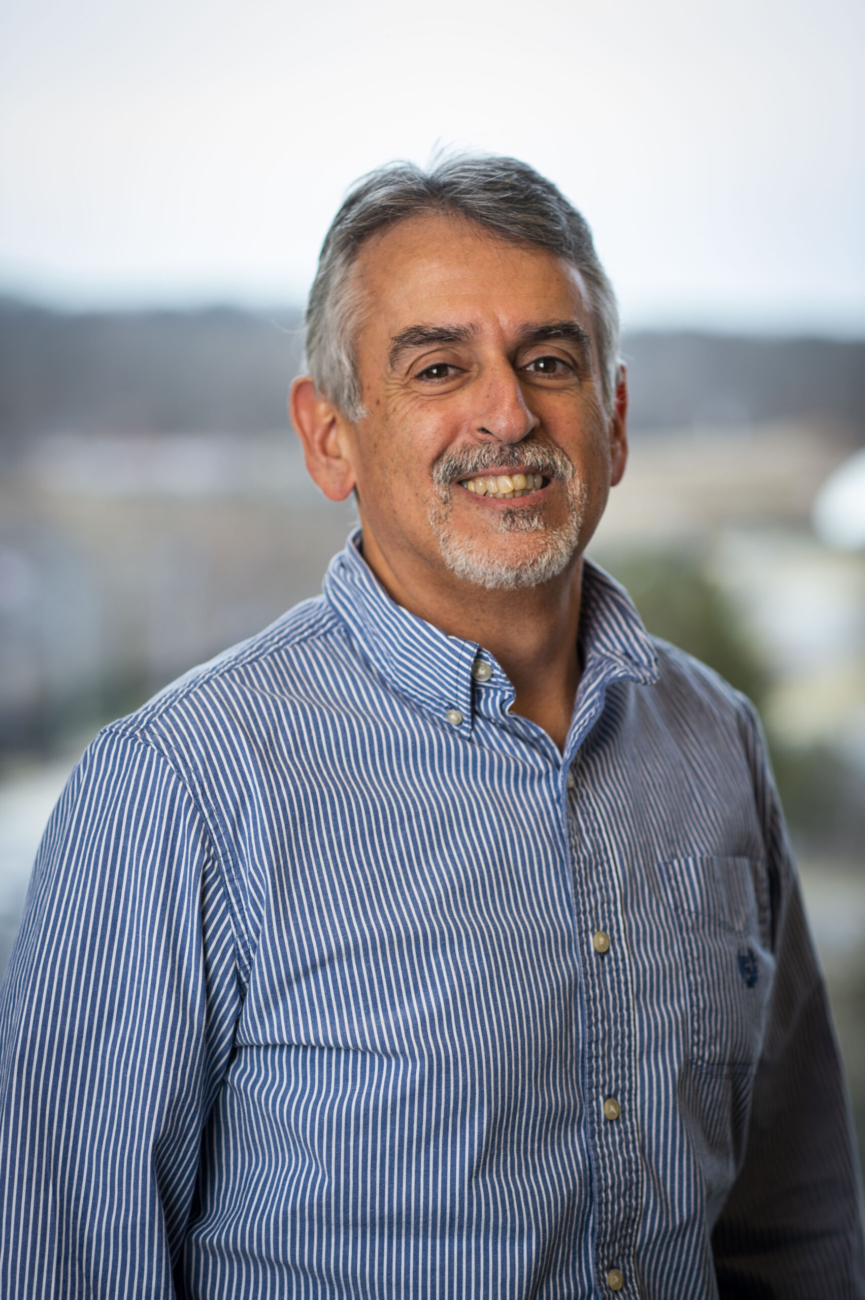 Ralph Ferraro wears a light blue plaid button-up, smiling in front of a blurred campus background