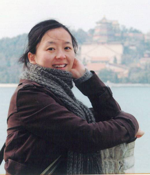 Dr. Yafang Cheng smiles in front of a watery vista, wearing a coat and scarf